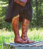 A person wearing the Bed|Stü Takoma brown leather boots with a western-shaped heel, paired with a black skirt and tan knee-high socks, stands on a wooden platform outdoors in a grassy area.