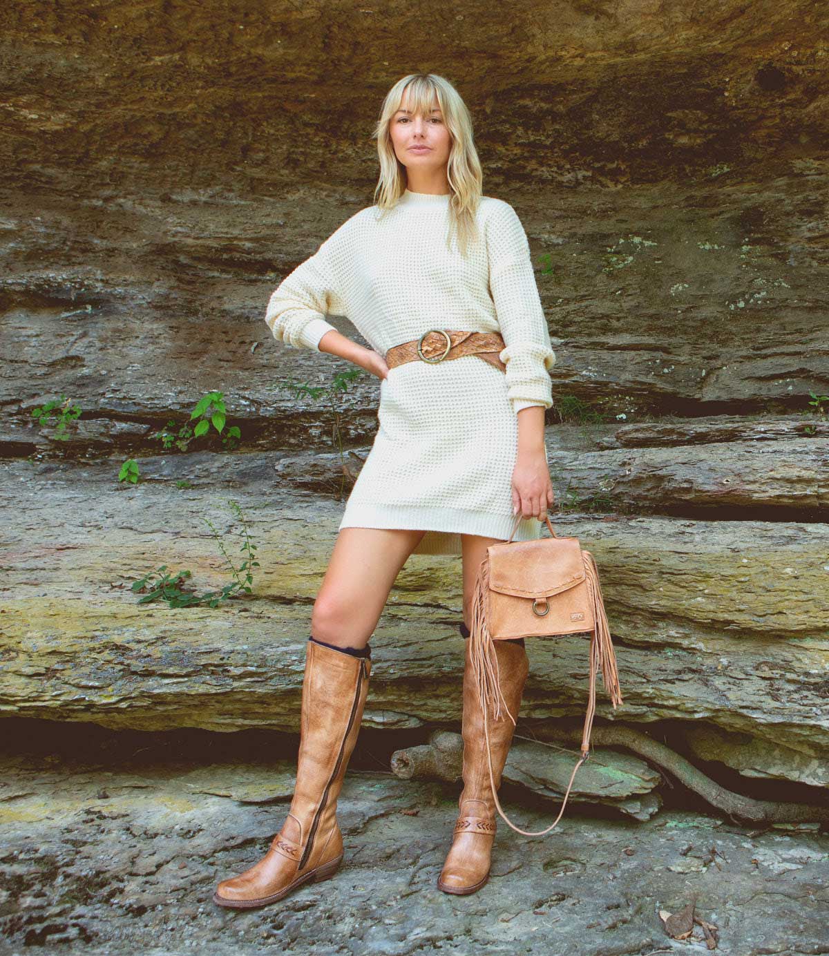 A person stands outdoors in front of rock layers, wearing a cream sweater dress, a brown belt, holding a brown fringe bag, and tan Bed|Stü Takoma knee-high boots with a western-shaped heel.