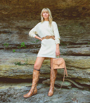 A person stands outdoors in front of rock layers, wearing a cream sweater dress, a brown belt, holding a brown fringe bag, and tan Bed|Stü Takoma knee-high boots with a western-shaped heel.
