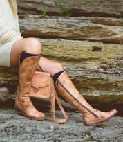 Wearing the Bed|Stü Takoma knee-high tan leather boots with a western-shaped heel, paired with a cream sweater and a matching tan purse while sitting on stone steps.