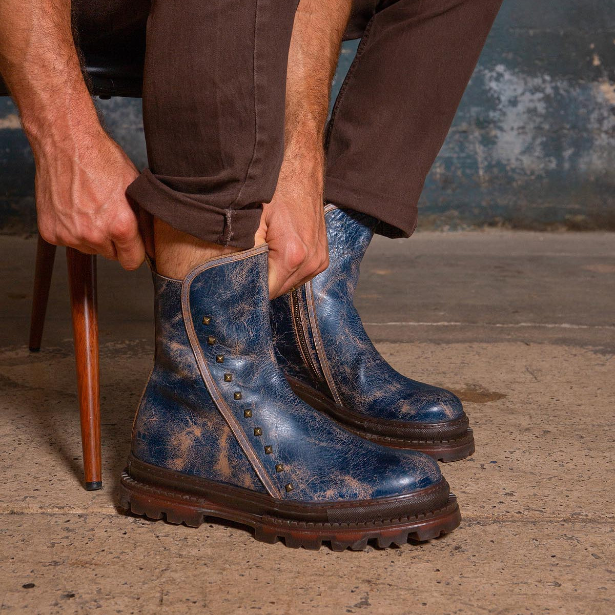 Seated on a chair against a worn concrete floor and textured wall, a person wearing brown pants puts on the Bed|Stü Tanto—distressed blue leather boots featuring metal studs and a rugged lug outsole.
