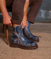 Seated on a chair against a worn concrete floor and textured wall, a person wearing brown pants puts on the Bed|Stü Tanto—distressed blue leather boots featuring metal studs and a rugged lug outsole.