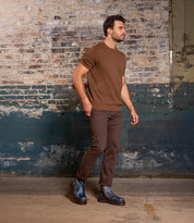 A man wearing a brown short-sleeve sweater, brown pants, and Bed|Stü's Tanto boots—featuring a leather upper and lug outsole—walks indoors against a worn brick wall.