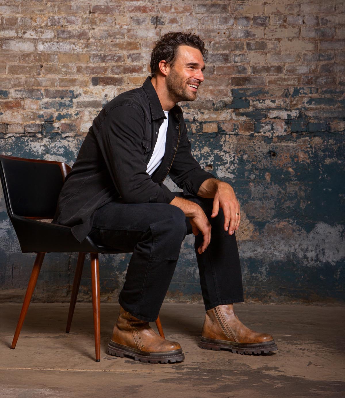 A man with dark hair and a beard sits sideways on a modern chair, smiling. He wears black attire and highlights the Tanto boot from Bed|Stü, which features a sleek leather upper and a lug outsole. The backdrop is a weathered, industrial brick wall.