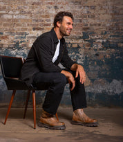 A man with dark hair and a beard sits sideways on a modern chair, smiling. He wears black attire and highlights the Tanto boot from Bed|Stü, which features a sleek leather upper and a lug outsole. The backdrop is a weathered, industrial brick wall.