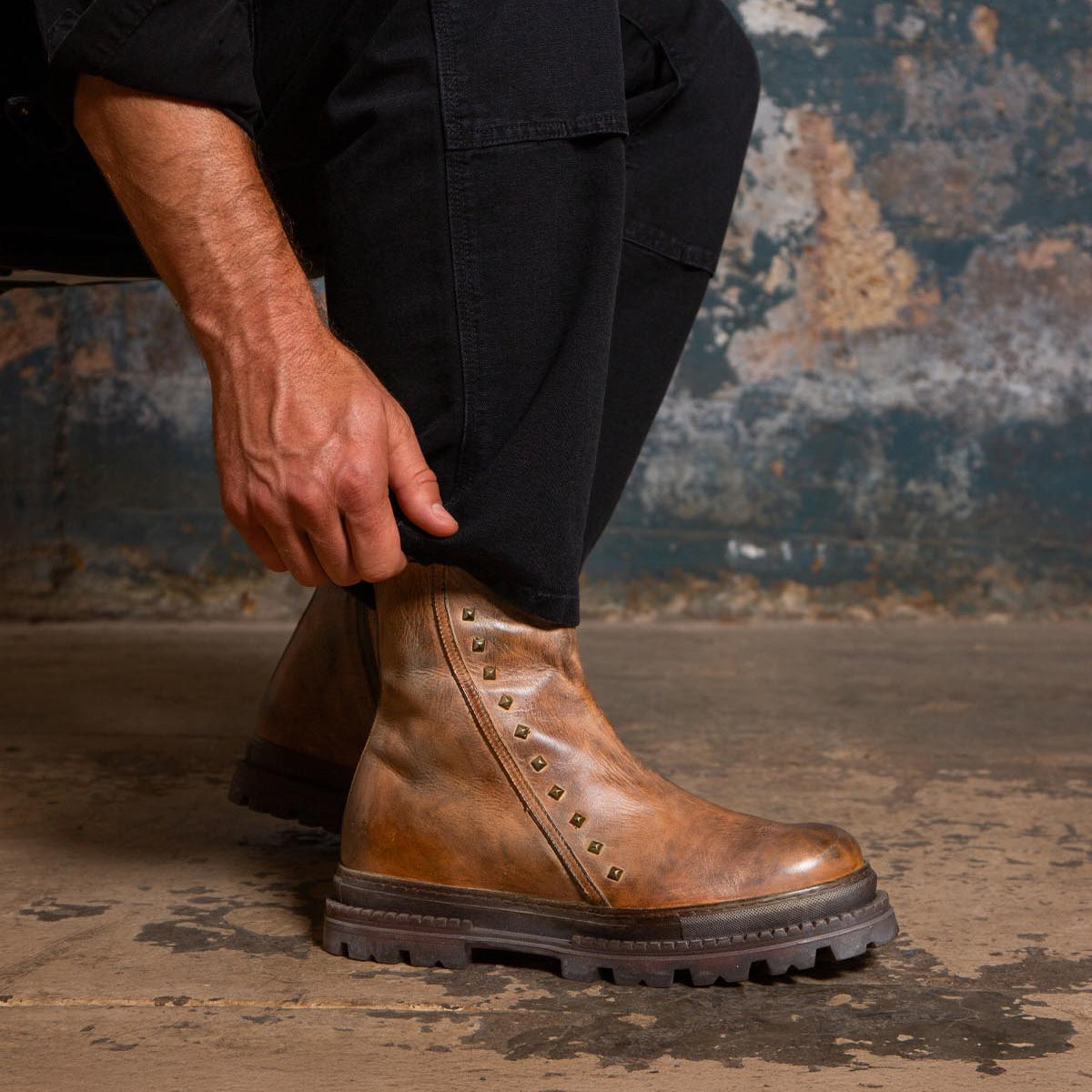 A person wearing black pants adjusts the Bed|Stü Tanto boot, featuring a streamlined design with a brown leather upper and metal studs, while sitting on a rough concrete floor against a distressed wall.