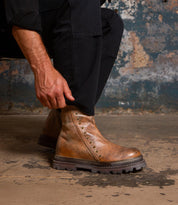 A person wearing black pants adjusts the Bed|Stü Tanto boot, featuring a streamlined design with a brown leather upper and metal studs, while sitting on a rough concrete floor against a distressed wall.