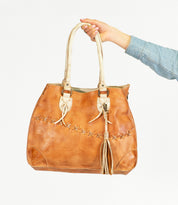 A hand holds the Bed|Stü Tassel Clip, a tan leather handbag featuring stitching details, dual handles, and a leather tassel accessory, against a white background.