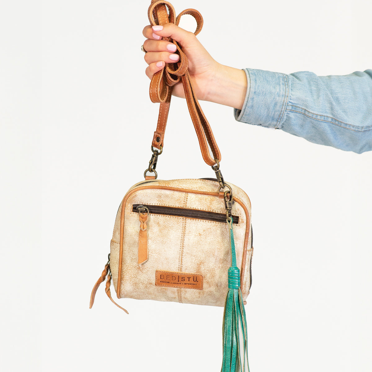A person in a denim sleeve holds a small tan leather crossbody bag with brown straps, accessorized with the Bed|Stü Tassel Clip in green leather, against a white background.