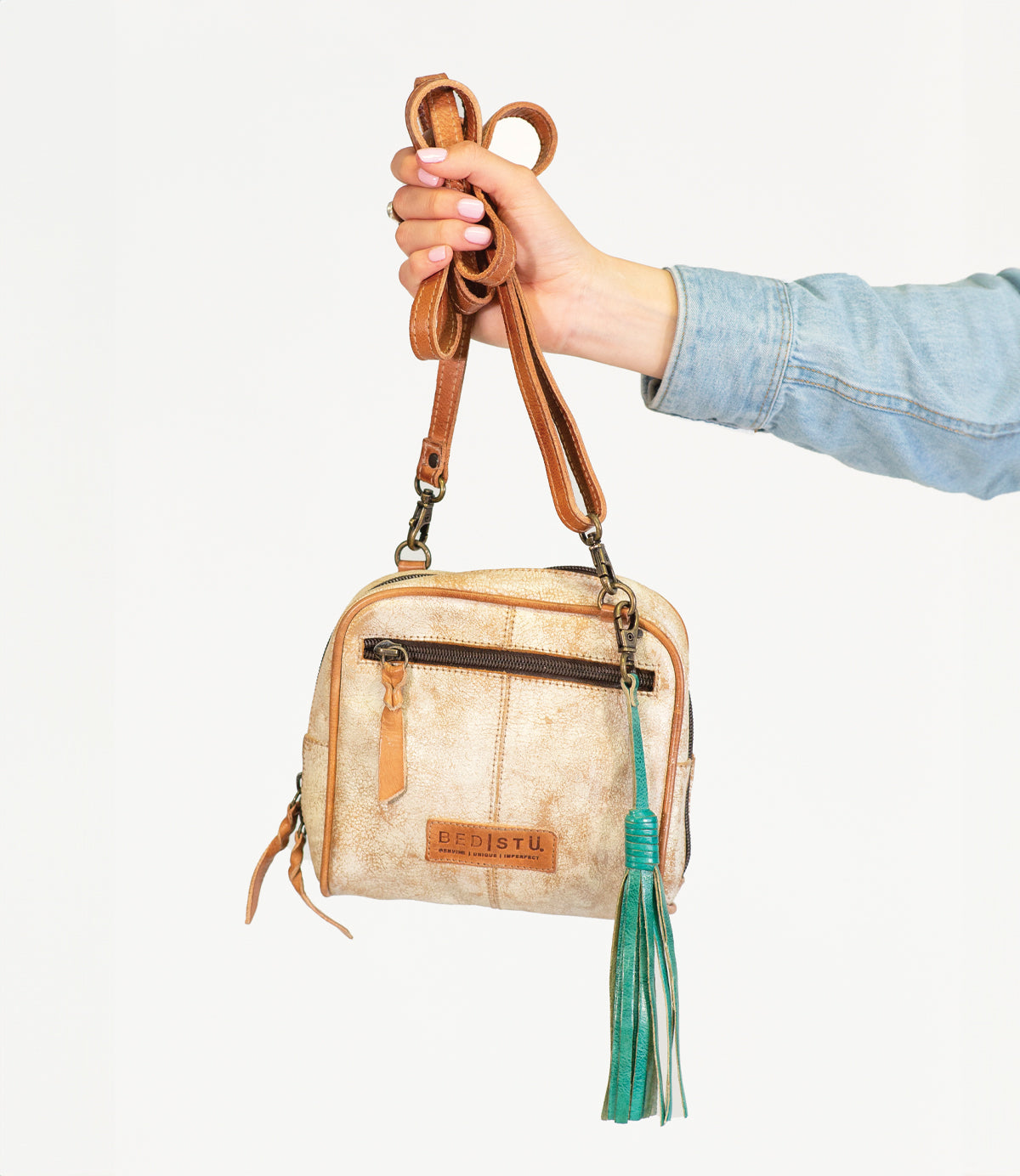 A person in a denim sleeve holds a small tan leather crossbody bag with brown straps, accessorized with the Bed|Stü Tassel Clip in green leather, against a white background.