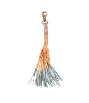 The Bed|Stü Tassel Clip features a leather keychain with a sturdy metal clasp and a long, multicolored fringe in light orange, beige, and blue tones—making it an eye-catching bag accessory or unique keychain charm.