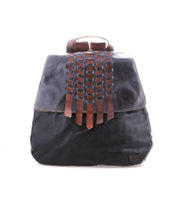The Teji backpack by Bed|Stü is crafted from black leather with a flap cover, featuring decorative handwoven brown leather accents on the front and a small logo patch in the lower corner, blending artisanal charm with timeless style.