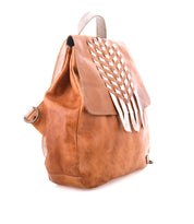 The Teji backpack by Bed|Stü is a brown handwoven leather bag featuring a flap top and a decorative white cutout pattern on the front, displayed at an angle against a white background.
