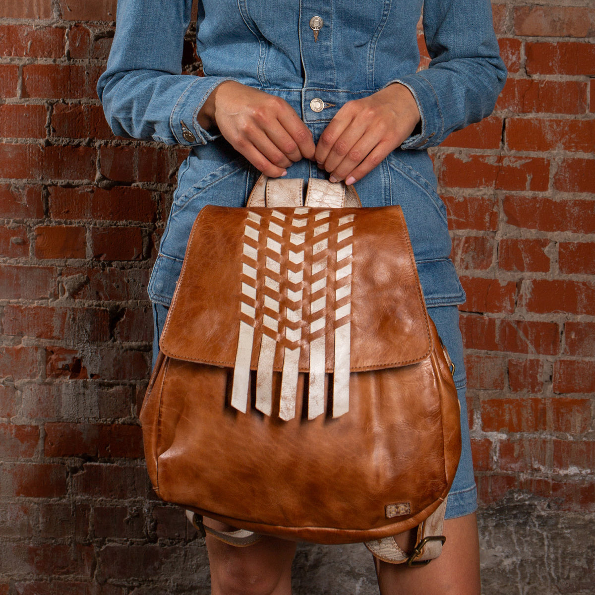 A person in a denim dress holds the Bed|Stü Teji handwoven leather bag, featuring white geometric accents, while standing against a red brick wall.