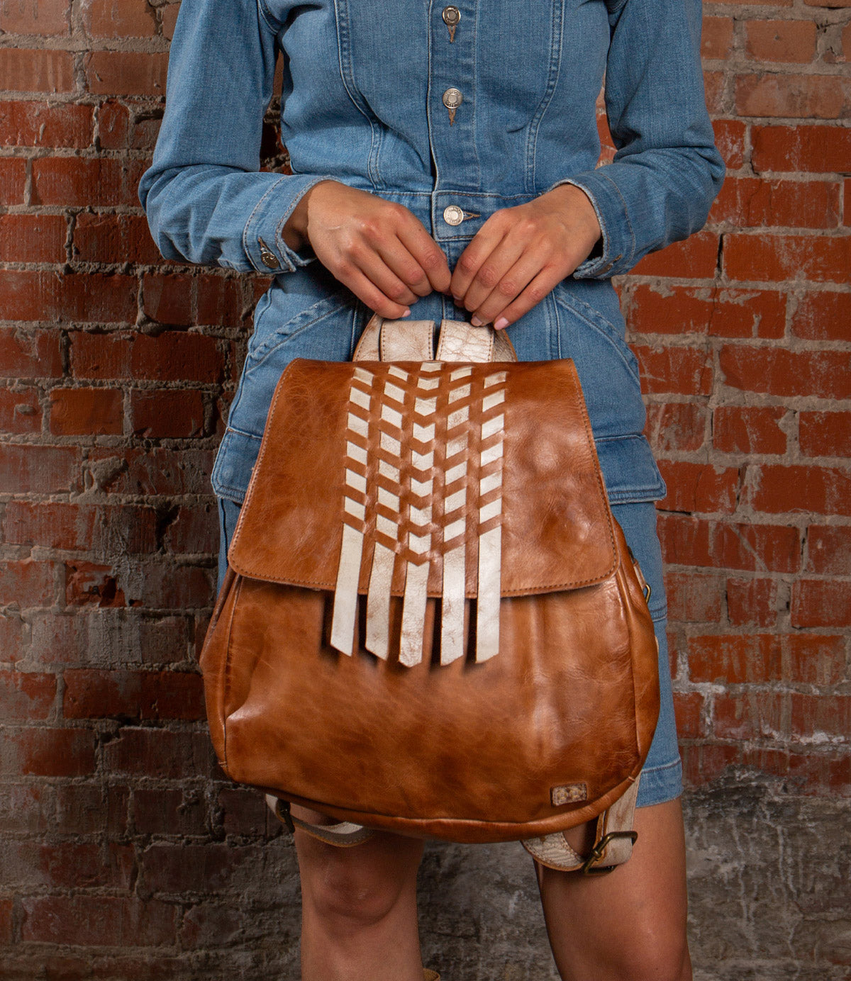 A person in a denim dress holds the Bed|Stü Teji handwoven leather bag, featuring white geometric accents, while standing against a red brick wall.