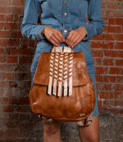 A person in a denim dress holds the Bed|Stü Teji handwoven leather bag, featuring white geometric accents, while standing against a red brick wall.