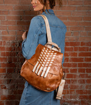 A woman in a denim shirt dress stands against a brick wall, carrying the Bed|Stü Teji backpack in brown leather with cream diagonal accents and an attached matching strap wallet. She completes her effortlessly chic look with this artisanal handwoven leather bag, adding a touch of distinctive charm to her ensemble.