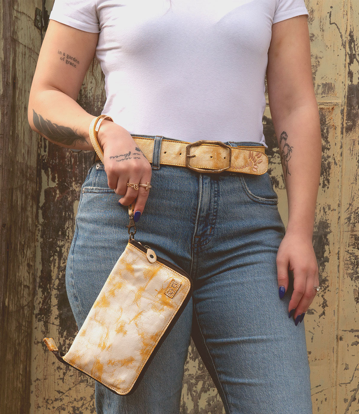 A person wearing a white t-shirt and blue jeans is holding the Bed|Stü Templeton II yellow tie-dye clutch wallet with a matching belt, standing in front of a weathered wooden wall.