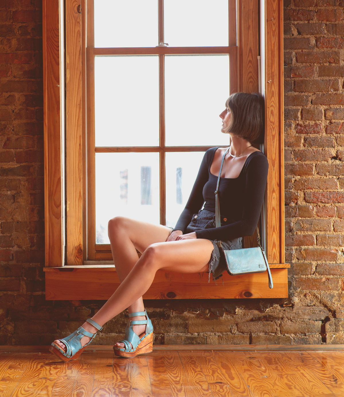 A woman with short dark hair sits on a wooden window ledge, wearing a black top, denim shorts, blue sandals, and carrying the Bed|Stü Templeton II sleek leather crossbody purse as she gazes out the window.