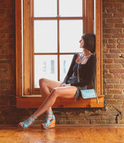 A woman with short dark hair sits on a wooden window ledge, wearing a black top, denim shorts, blue sandals, and carrying the Bed|Stü Templeton II sleek leather crossbody purse as she gazes out the window.