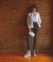 A woman stands against a brick wall, wearing a white jacket, crop top, dark jeans, gray ankle boots, and carrying the Bed|Stü Templeton II sleek leather crossbody.