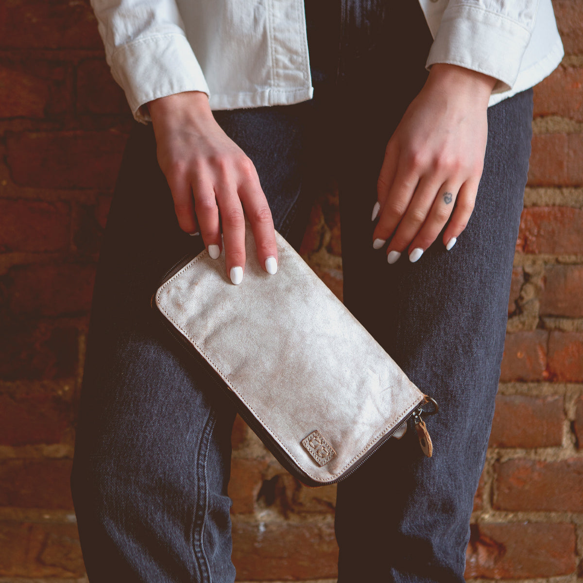 A person wearing a white shirt and dark jeans holds the Bed|Stü Templeton II metallic clutch purse with both hands in front of a brick wall, effortlessly showcasing their chic style with this elegant accessory.