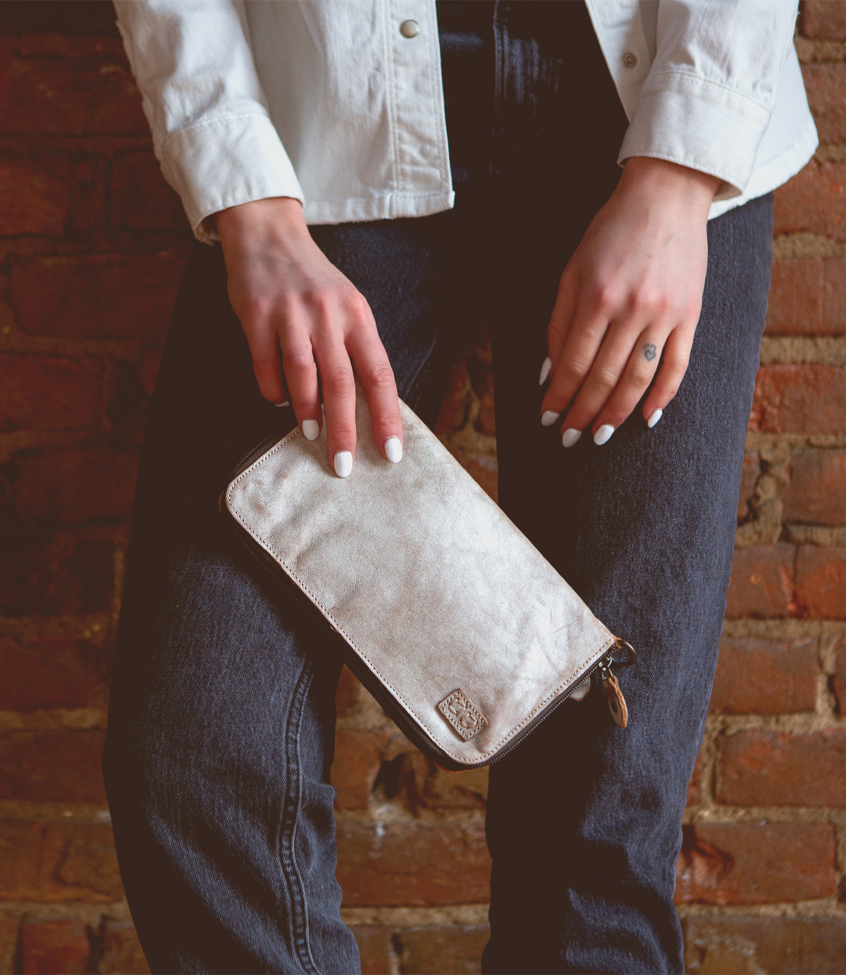 A person wearing a white shirt and dark jeans holds the Bed|Stü Templeton II metallic clutch purse with both hands in front of a brick wall, effortlessly showcasing their chic style with this elegant accessory.