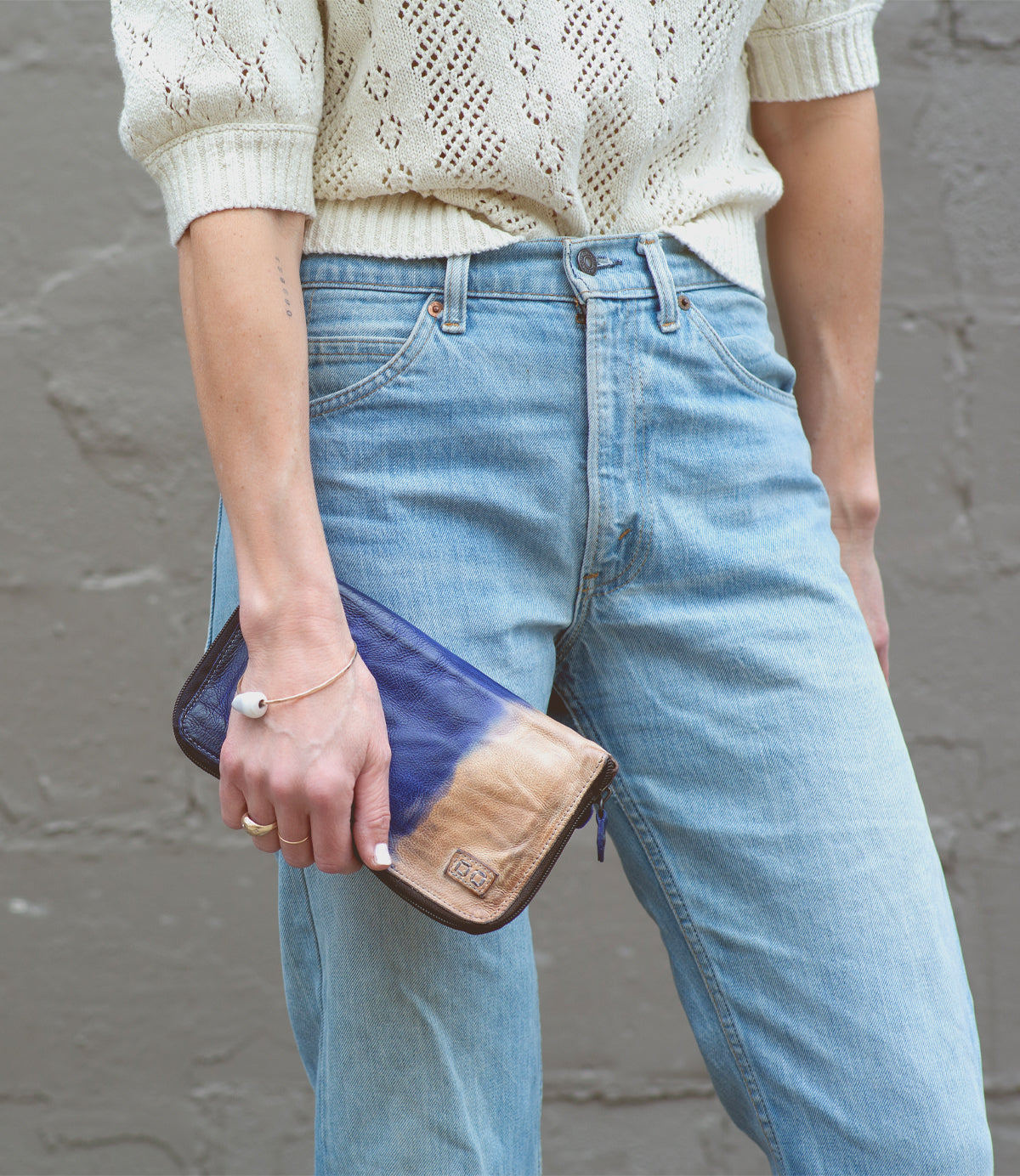 A person wearing a cream knit top and light blue jeans holds the Bed|Stü Templeton II wallet clutch in blue and tan against a gray textured wall.