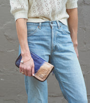 A person wearing a cream knit top and light blue jeans holds the Bed|Stü Templeton II wallet clutch in blue and tan against a gray textured wall.