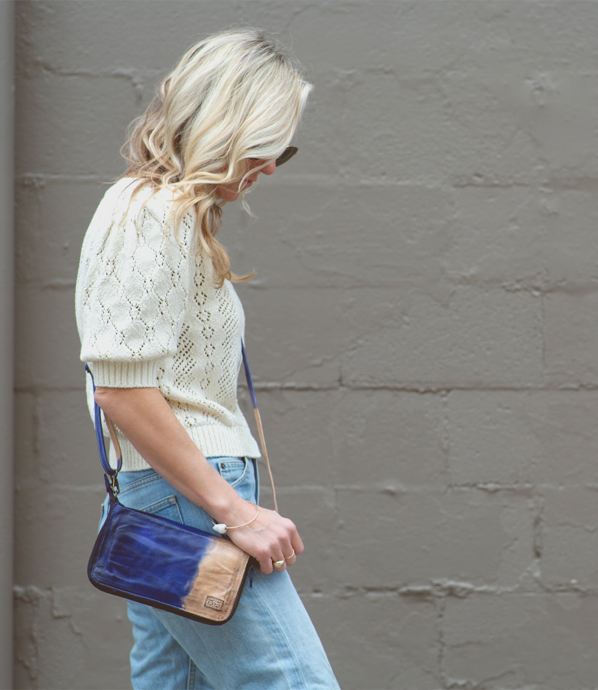 A woman with wavy blonde hair, dressed in a white knit top and blue jeans, walks past a textured gray wall while carrying the Bed|Stü Templeton II—a stylish leather crossbody bag in blue and tan.