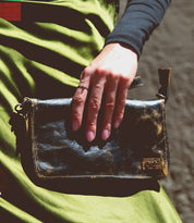 A hand adorned with the Bed|Stü Templeton II ring rests on a shiny black clutch, set against a backdrop of green satin fabric.