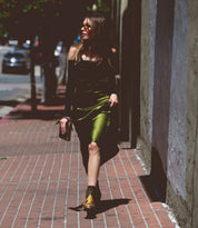 A woman in a green dress and black boots walks on a sunlit city sidewalk, holding the Templeton II small leather crossbody clutch by Bed|Stü.