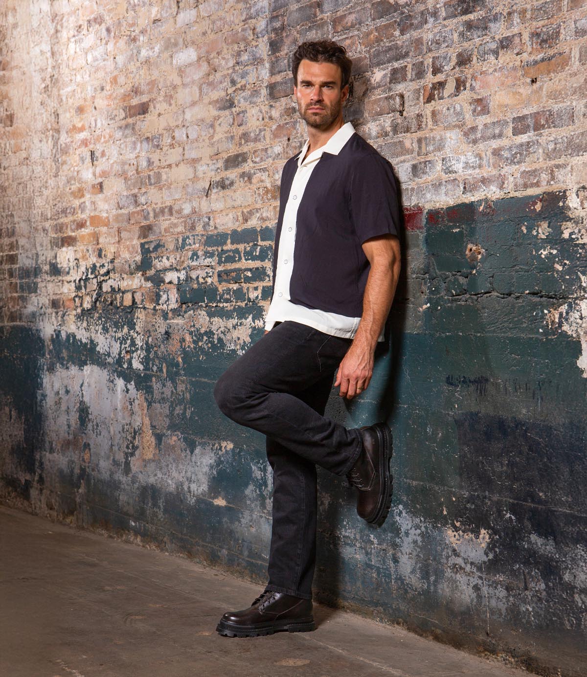 A man stands against a distressed brick wall with one leg bent, wearing a short-sleeve shirt over a white top, black pants, and a pair of Bed|Stü Tempt Chelsea boots for a stylish hybrid look.