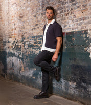 A man stands against a distressed brick wall with one leg bent, wearing a short-sleeve shirt over a white top, black pants, and a pair of Bed|Stü Tempt Chelsea boots for a stylish hybrid look.