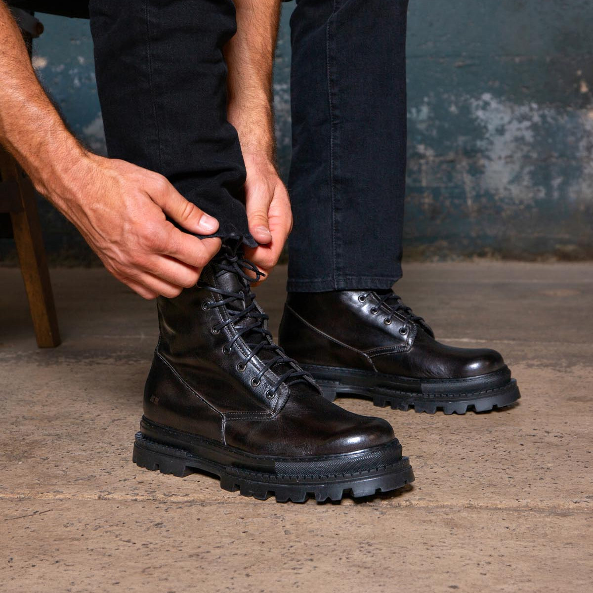 A person wearing black jeans ties the laces of Bed|Stü Tempt hybrid black leather lace-up boots while sitting on a chair, with a worn blue wall and concrete floor in the background.