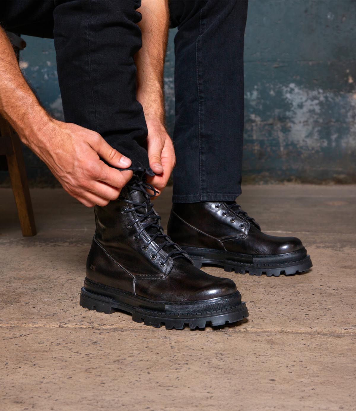 A person wearing black jeans ties the laces of Bed|Stü Tempt hybrid black leather lace-up boots while sitting on a chair, with a worn blue wall and concrete floor in the background.
