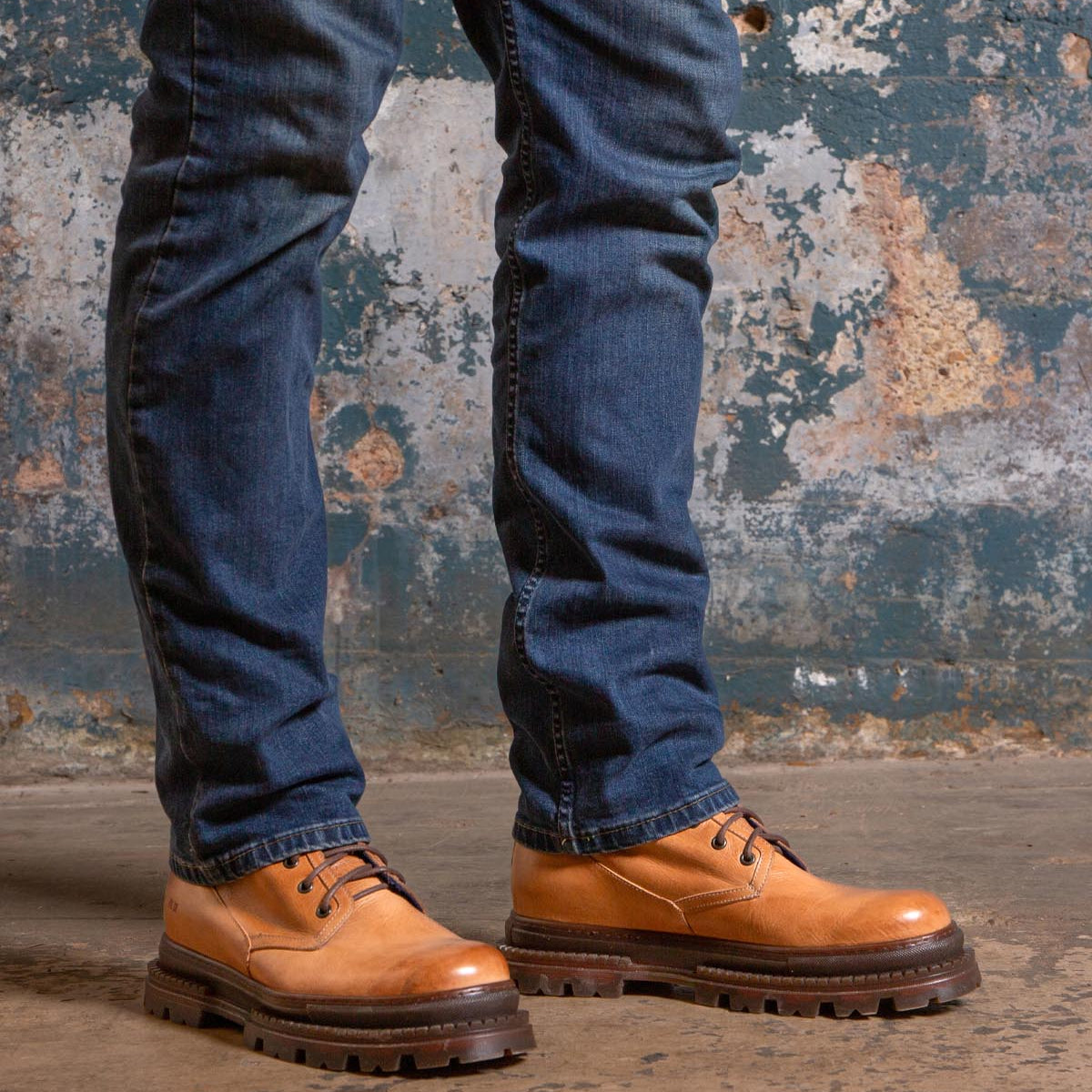 A person wearing blue jeans and Bed|Stü’s Tempt tan lace-up leather shoes—designed as a stylish hybrid between a hiking boot and Chelsea boot—stands on a concrete floor against a textured, weathered wall.