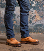 A person wearing blue jeans and Bed|Stü’s Tempt tan lace-up leather shoes—designed as a stylish hybrid between a hiking boot and Chelsea boot—stands on a concrete floor against a textured, weathered wall.