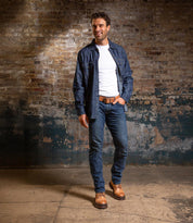 A man stands indoors against a textured brick wall, wearing a white t-shirt, an open denim shirt, jeans, and the Bed|Stü Tempt hybrid hiking boots, looking slightly to the side with a smile.