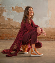 A woman wearing the Bed|Stü Twist tan high top sneakers with sleek leather panels kneels on one knee indoors, dressed in a maroon outfit and posed against a worn, textured wall.