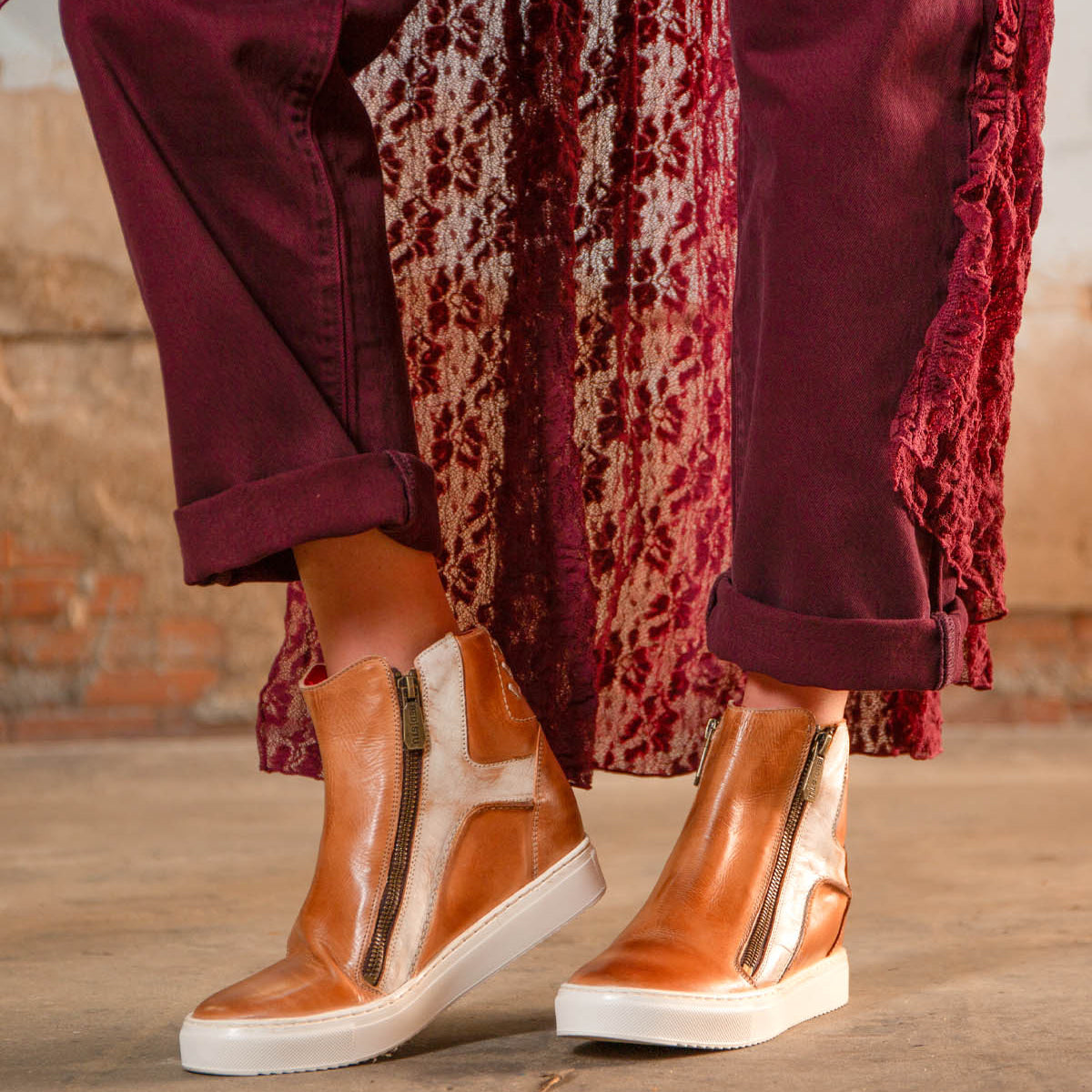 A person wearing the Bed|Stü Twist brown leather high-top sneakers with leather panels and side zippers, burgundy pants, and a patterned burgundy coat, standing on a concrete floor.