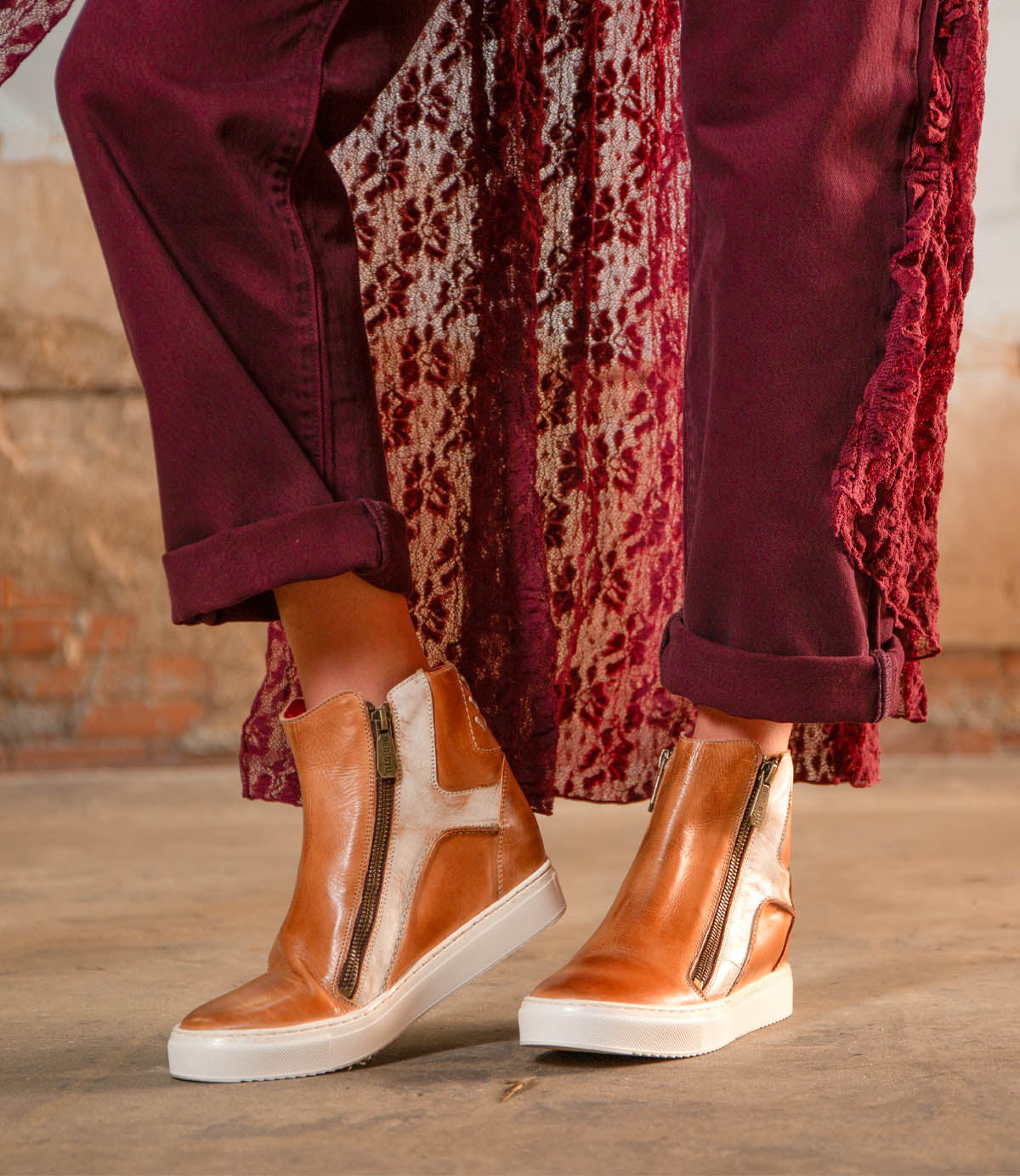 A person wearing the Bed|Stü Twist brown leather high-top sneakers with leather panels and side zippers, burgundy pants, and a patterned burgundy coat, standing on a concrete floor.