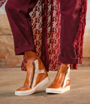 A person wearing the Bed|Stü Twist brown leather high-top sneakers with leather panels and side zippers, burgundy pants, and a patterned burgundy coat, standing on a concrete floor.