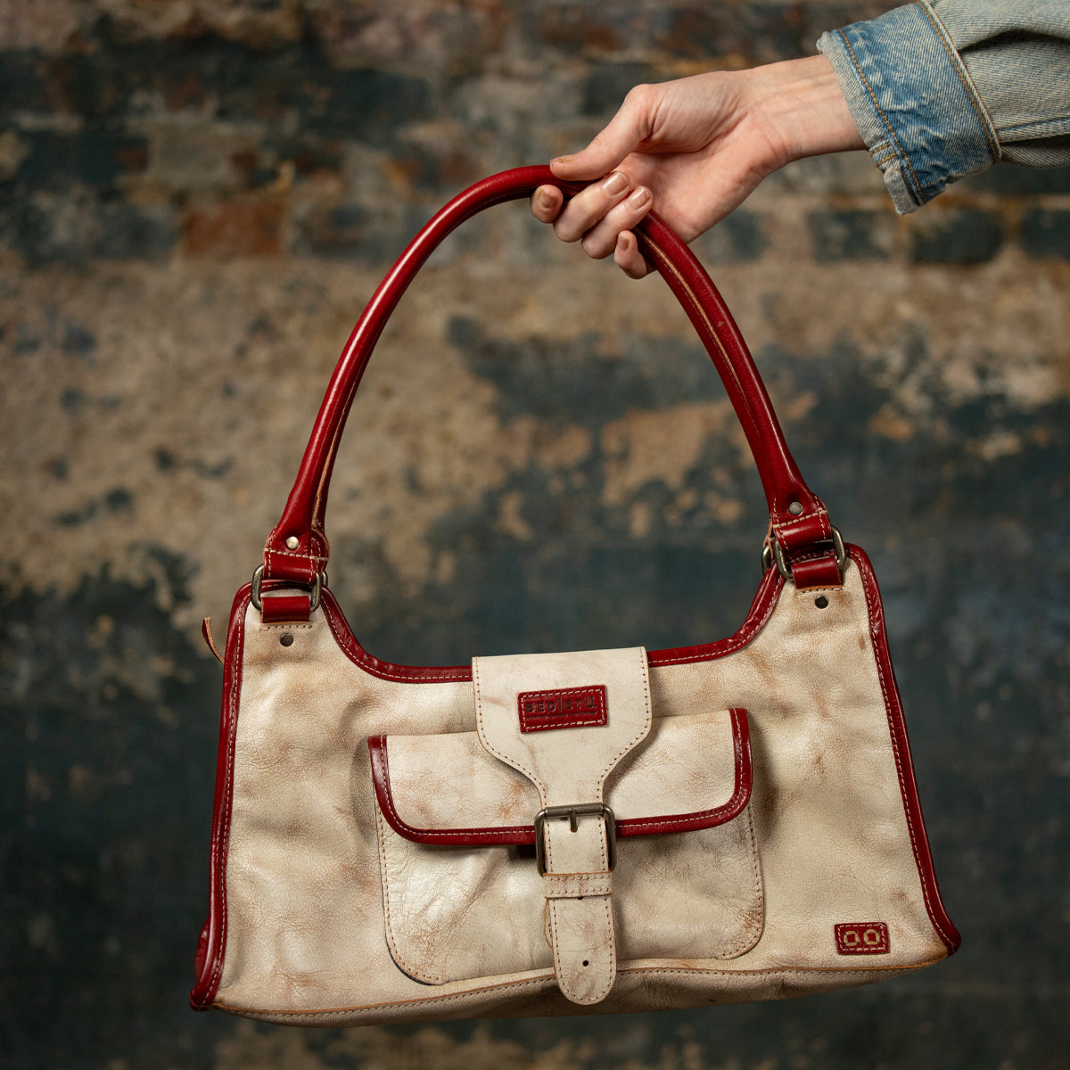 A hand holds the Bed|Stü "Unbending" washed leather handbag in beige, detailed with red handles and trim and a distinctive front buckle pocket, set against a textured, worn wall—the perfect elegant shoulder bag or versatile work-bag.