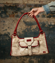 A hand holds the Bed|Stü "Unbending" washed leather handbag in beige, detailed with red handles and trim and a distinctive front buckle pocket, set against a textured, worn wall—the perfect elegant shoulder bag or versatile work-bag.