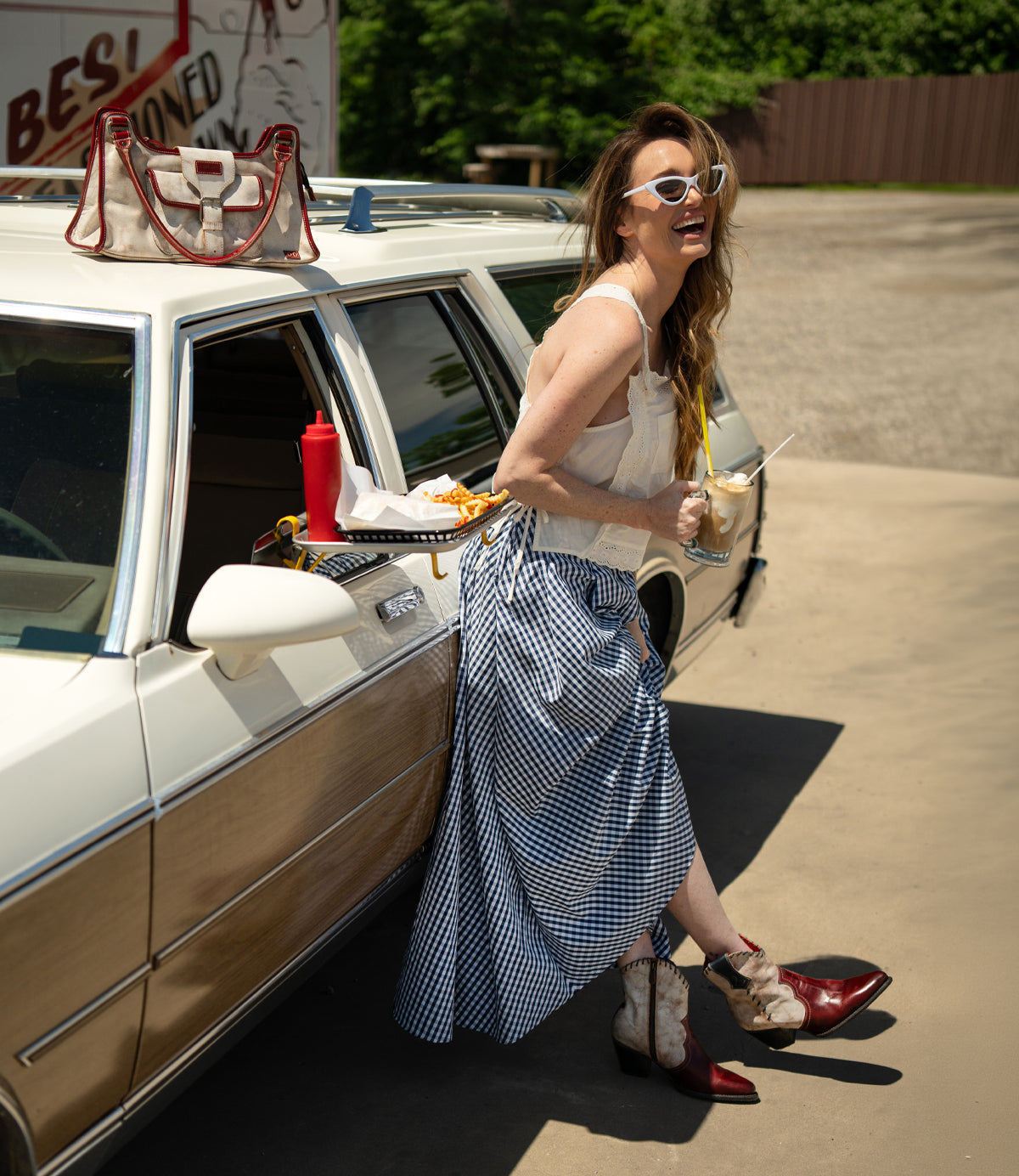 A woman in sunglasses stands beside a vintage car, holding a drink and fries. A tray with food is attached to the car door. She wears a blue gingham skirt, a white top, and carries the Bed|Stü Unbending washed leather shoulder bag, adding effortless style to her retro look.
