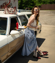 A woman in sunglasses stands beside a vintage car, holding a drink and fries. A tray with food is attached to the car door. She wears a blue gingham skirt, a white top, and carries the Bed|Stü Unbending washed leather shoulder bag, adding effortless style to her retro look.