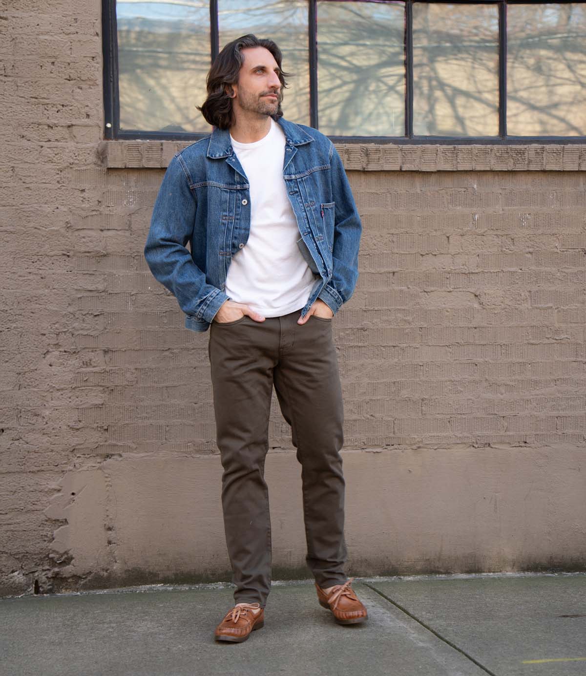 A man stands on a sidewalk in front of a beige brick wall with a large window, wearing a denim jacket, white t-shirt, dark pants, and Bed|Stü’s Uncle Floyd classic brown boat shoes featuring moc-toe stitching.