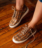 A person wearing Bed|Stü Unwavering handwoven leather sneakers with white soles stands on a wooden floor.
