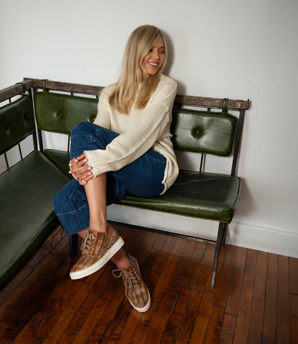 A woman with long blonde hair sits on a green bench, wearing a cream sweater, blue jeans, and the Unwavering sneakers by Bed|Stü, smiling and looking to the side.